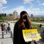 A person wearing a mask and a black beanie holds a yellow sign with the words 