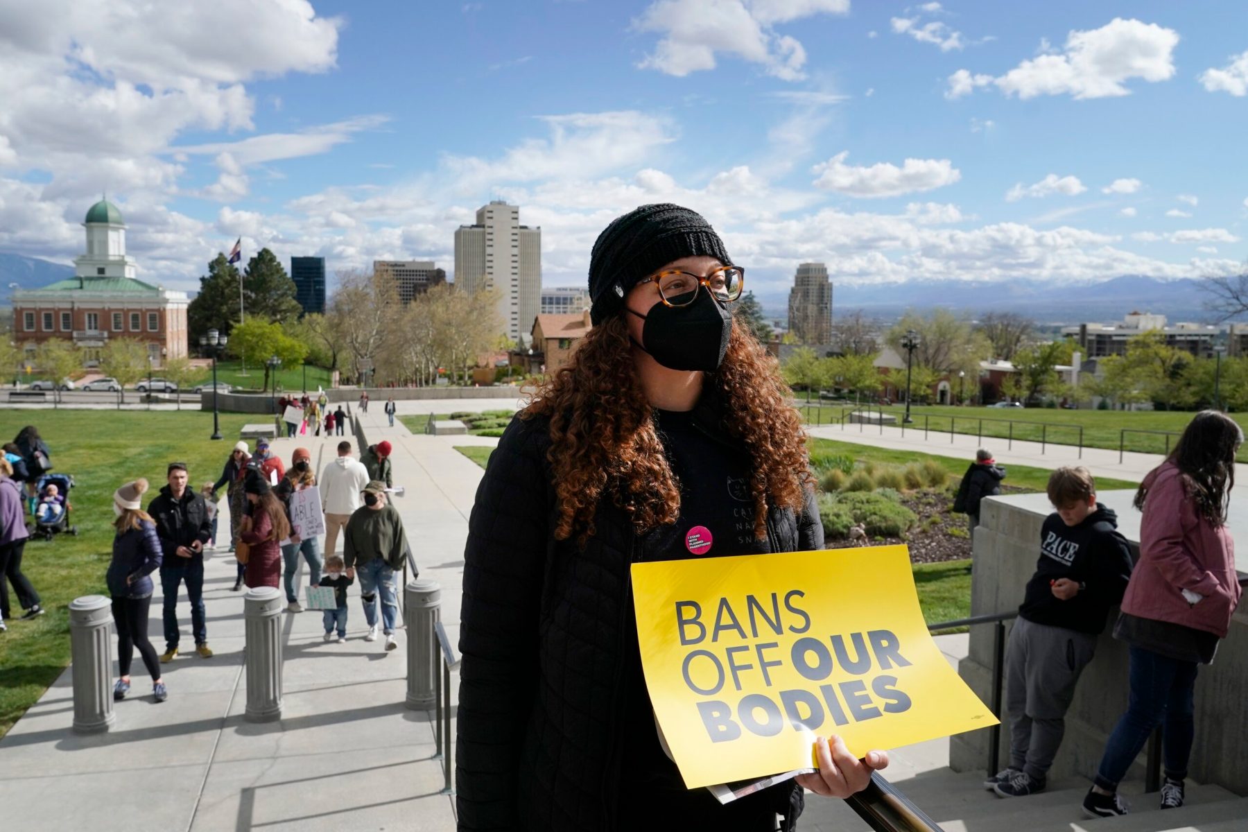 A person wearing a mask and a black beanie holds a yellow sign with the words "Bans off our bodies" on it.