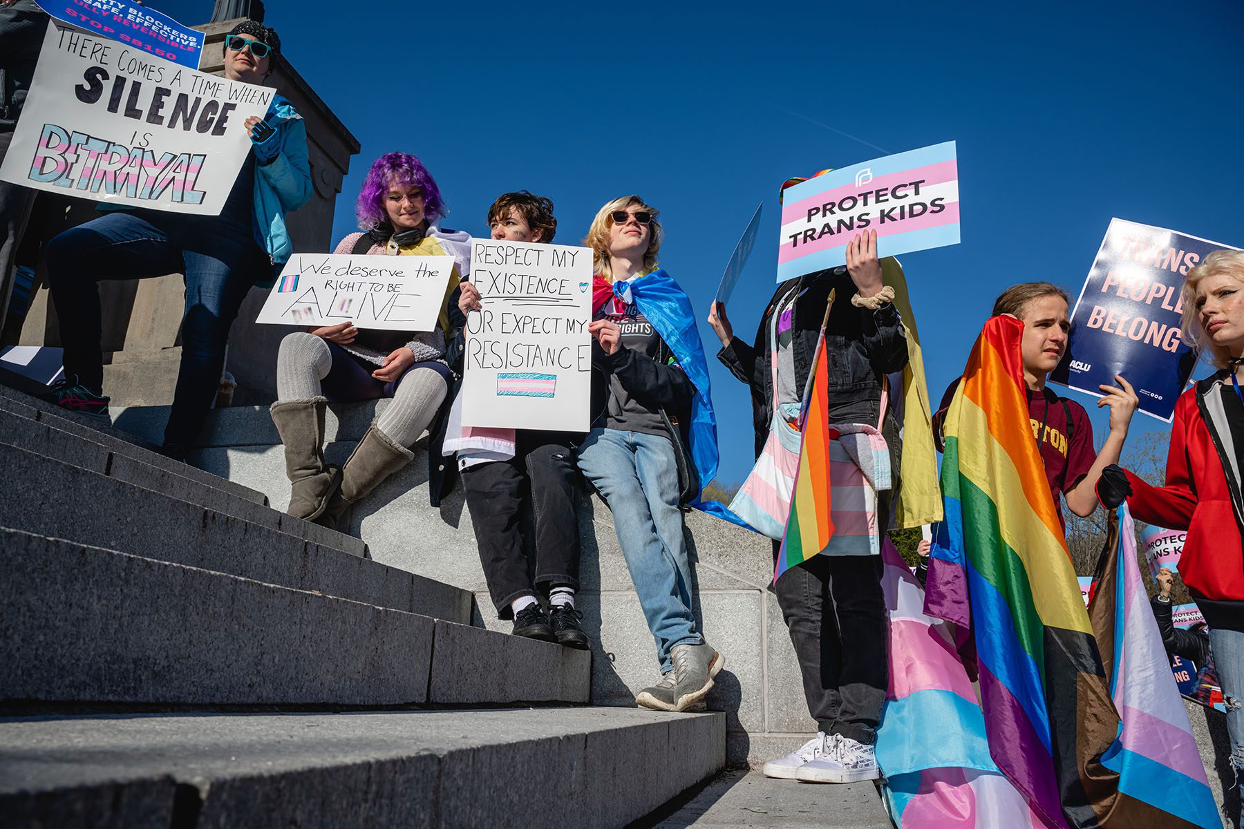Demonstrators gather at a rally to protest the passing of anti-trans bills at the Kentucky State Capitol. They hold signs that read "We deserve to be alive," "There comes a time when silence is betrayal," "protect trans kids," and "Respect my existence or expect my resistance."