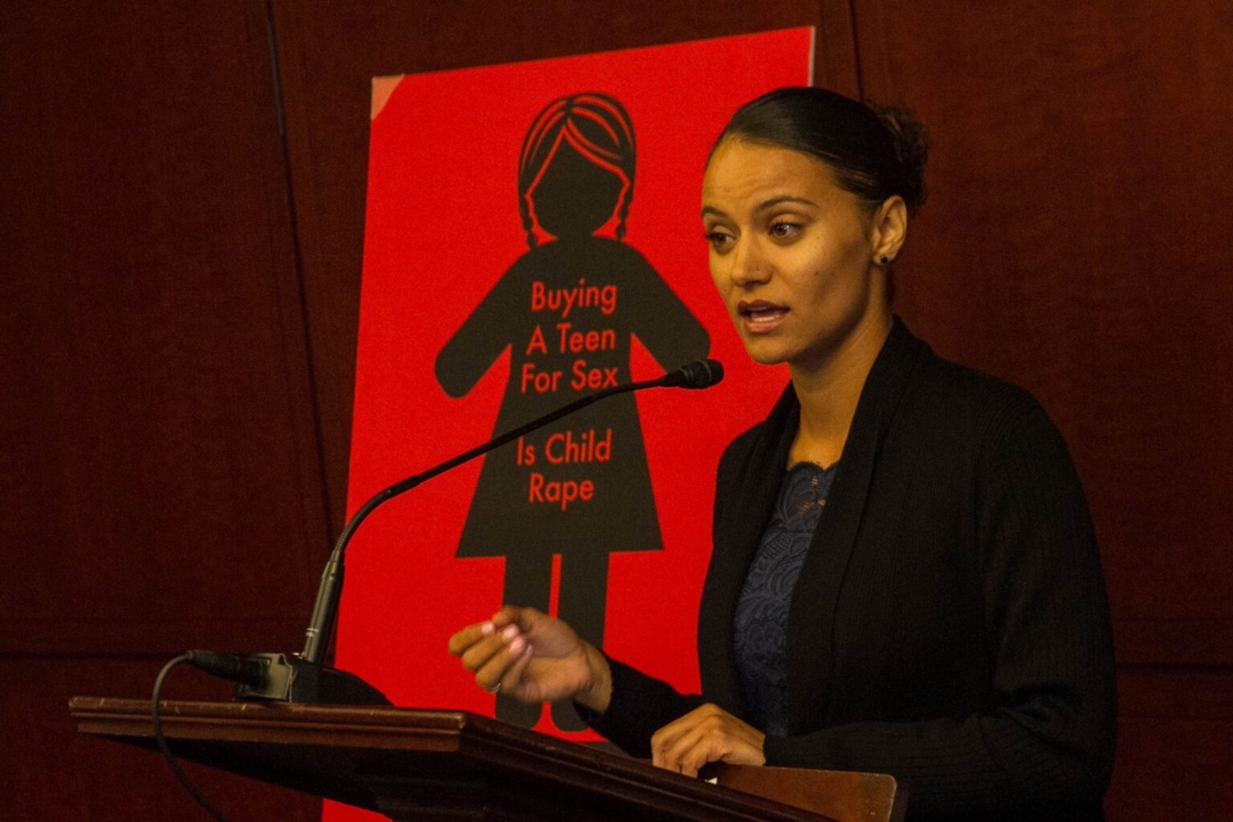 T Ortiz stands at a podium speaking into a microphone. Behind her is a red poster that reads, "buying a teen for sex is child rape."