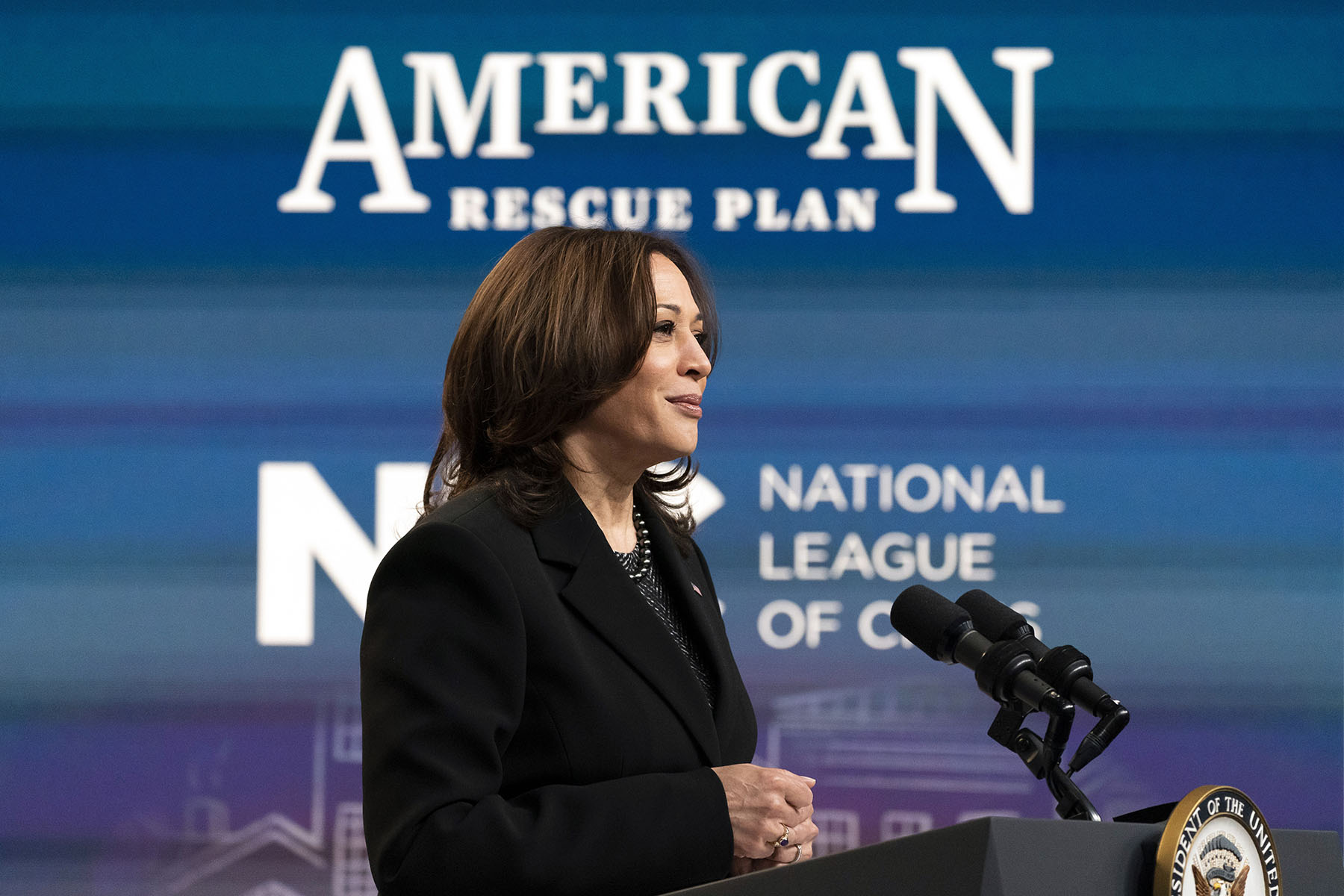 Vice President Kamala Harris speaks at a podium on the White House campus while American Rescue Plan signage is seen behind her.