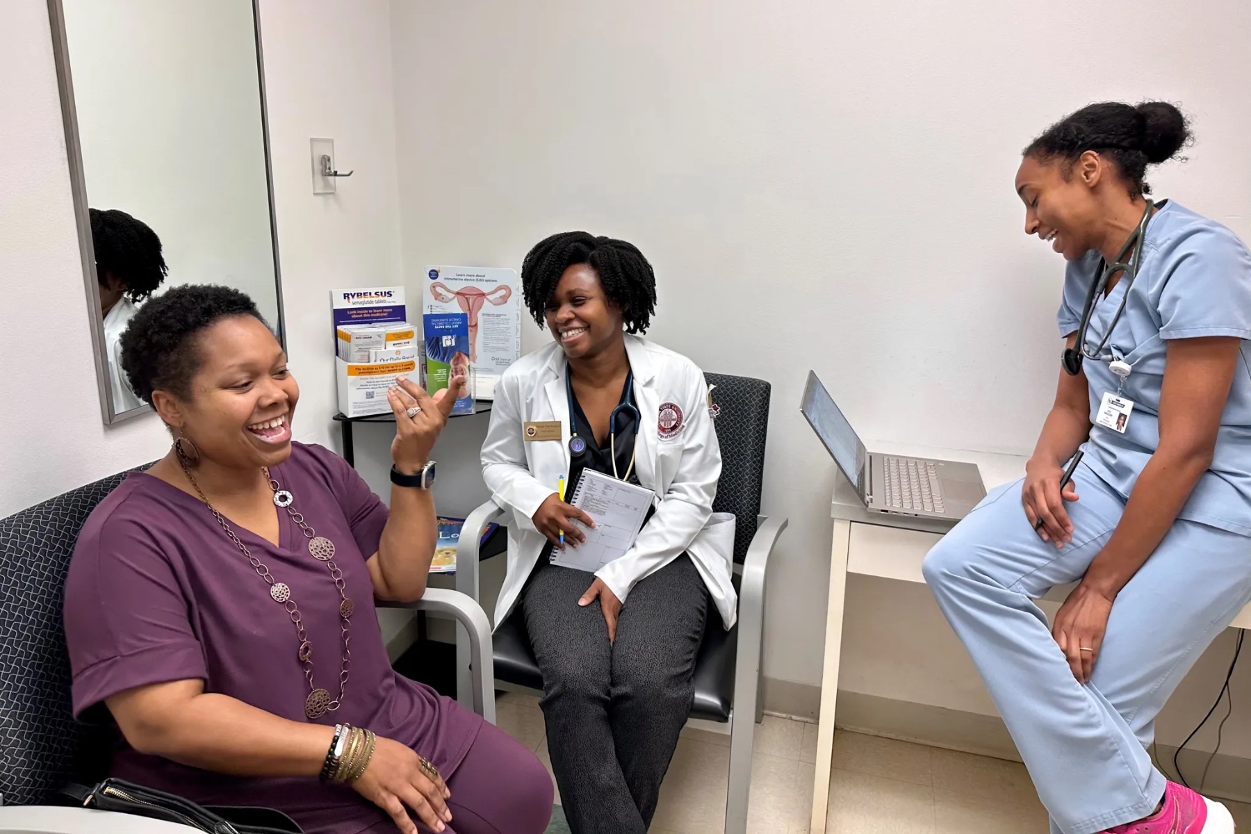 A smiling woman gestures at two medical professionals who are also smiling.