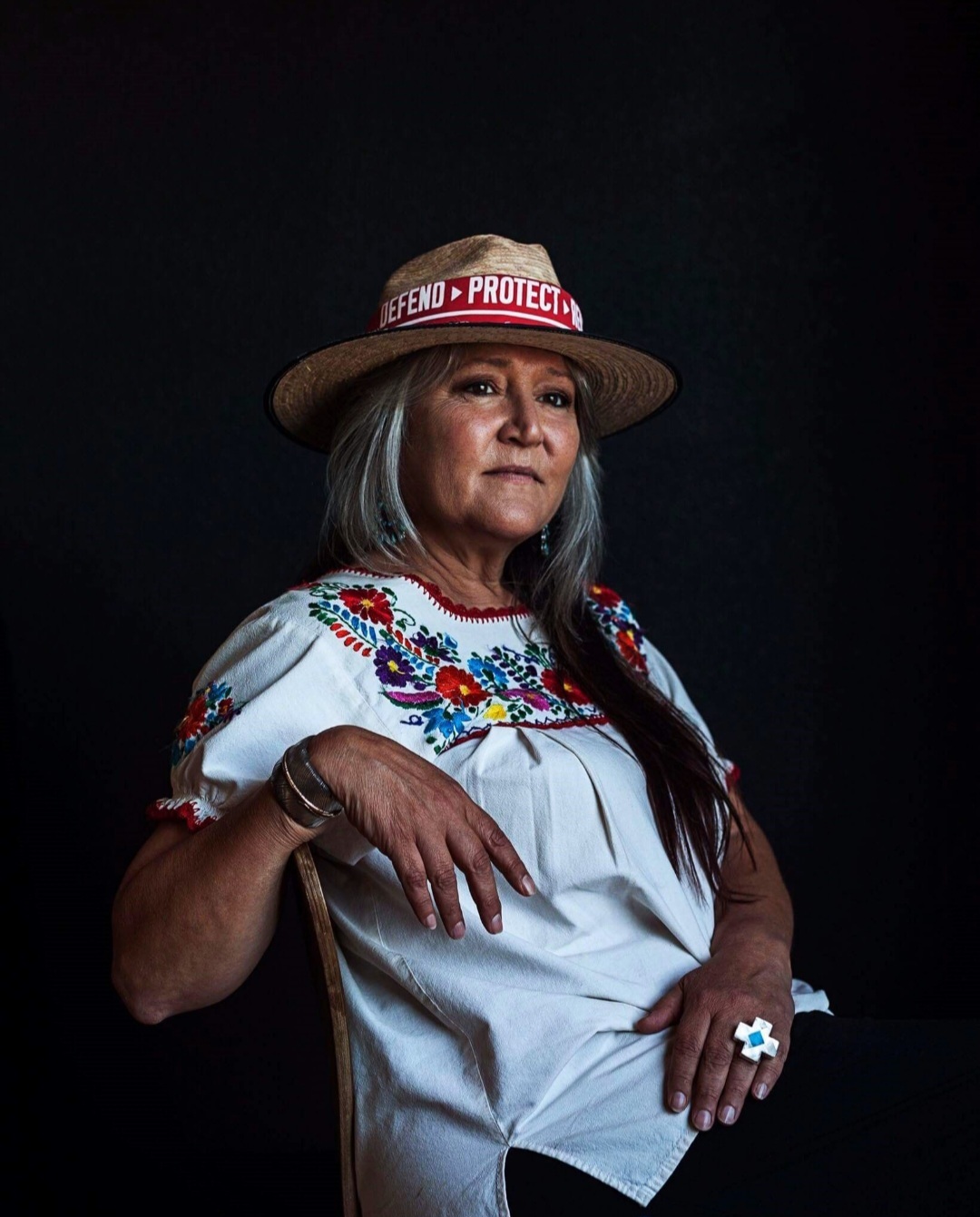 Pennie Opal Plant poses for a portrait. She sits in a chair and wears a hat that reads "Defend, Protect."