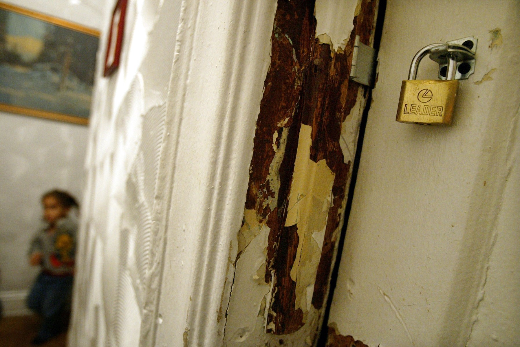 A child stands in the background near a wall of peeling paint. A doorway of peeling paint is in the foreground.