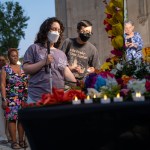 A woman holding a cane and a man in glasses, both wearing masks, are at the head of a line to put candles on a flower-covered memorial table in New York.