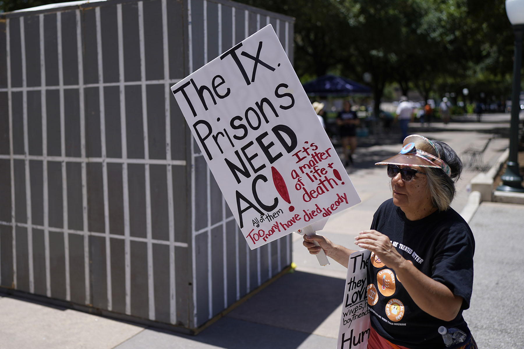 An advocate for cooling Texas prisons walks past a makeshift cell during a rally on the steps of the Texas Capitol. She is holding a sign that reads "The Tx. Prisons NEED AC! All of them. It's a matter of life and death! Too many have died already."