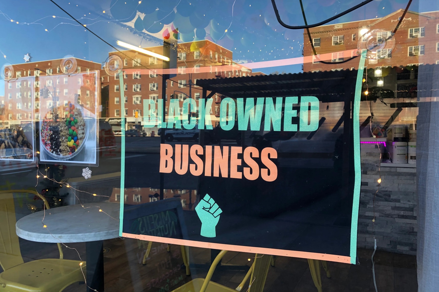 A black poster, reading "Black-owned business" with a raised fist beneath the text, hangs in a store window in Queens, New York. Low-rise red-brick apartments are reflected in the window.