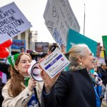 Demonstrators outside the Supreme Court; one holds a box of mifepristone tablets, one with a bullhorn, one with a sign that says 