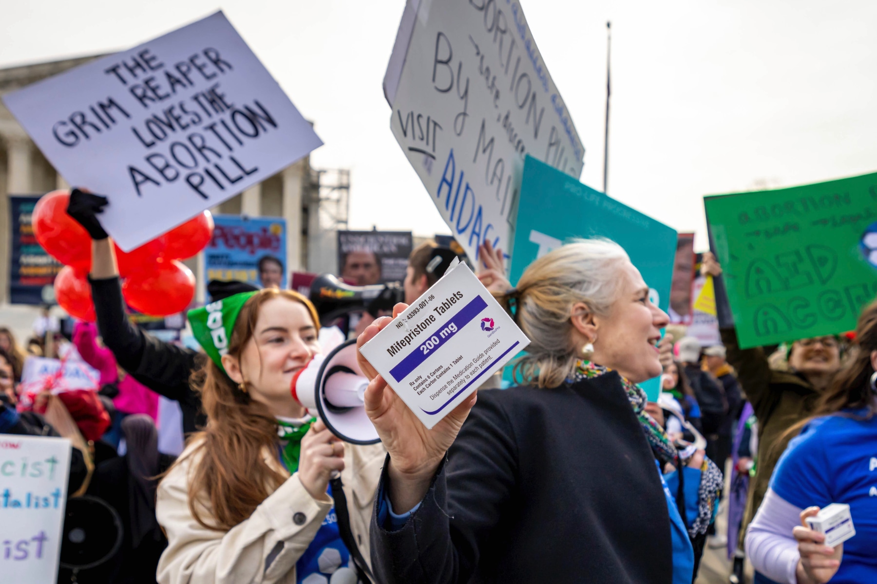 Demonstrators outside the Supreme Court; one holds a box of mifepristone tablets, one with a bullhorn, one with a sign that says "The grim reaper loves the abortion pill" and some with Aid Access signs