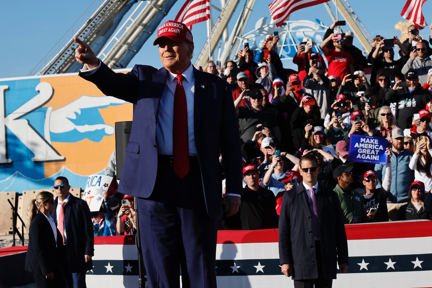 Donald Trump, wearing a suit and a re MAGA hat, points at a crowd at a rally in New Jersey