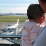 A mother holds her baby at the airport. In the background, the tarmac is seen through a window.