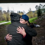 Unhoused folks hug each other under a bridge.