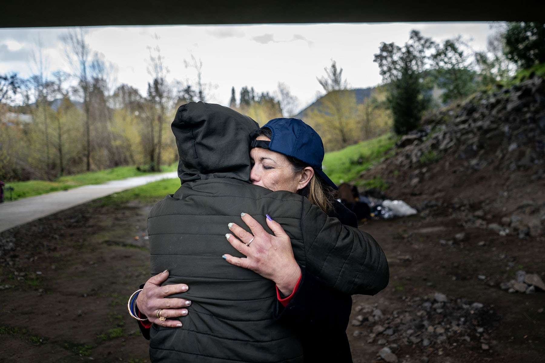 Unhoused folks hug each other under a bridge.