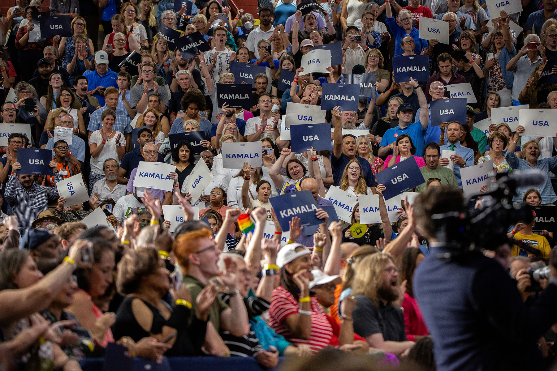 Supporters of VP Kamala Harris hold dark-blue-and-white signs that read “Kamala” on one side and “USA” on the other as they cheer during a campaign rally.