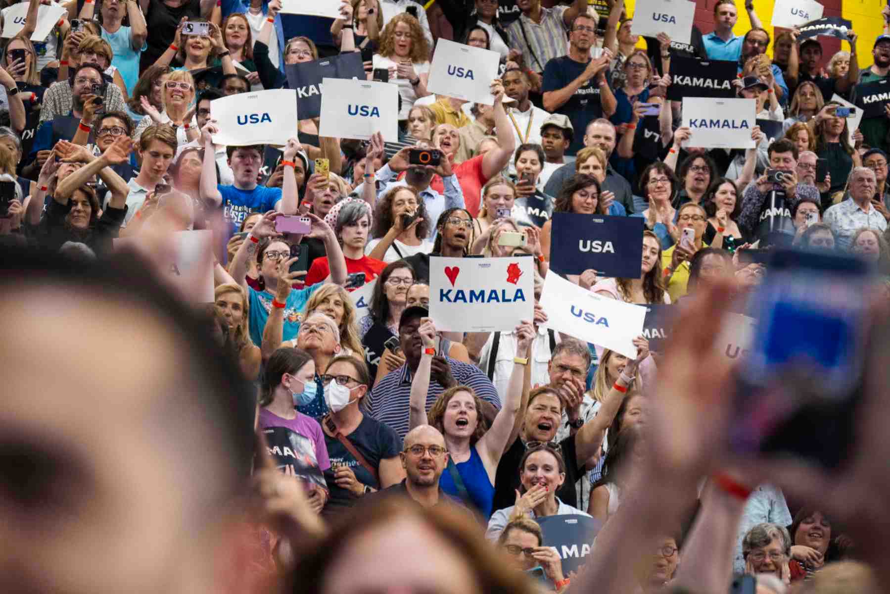 A large crowd of people attend a camapaign event for Vice President Kamala Harris. Several hold up signs that read "USA" and others that read "Kamala."