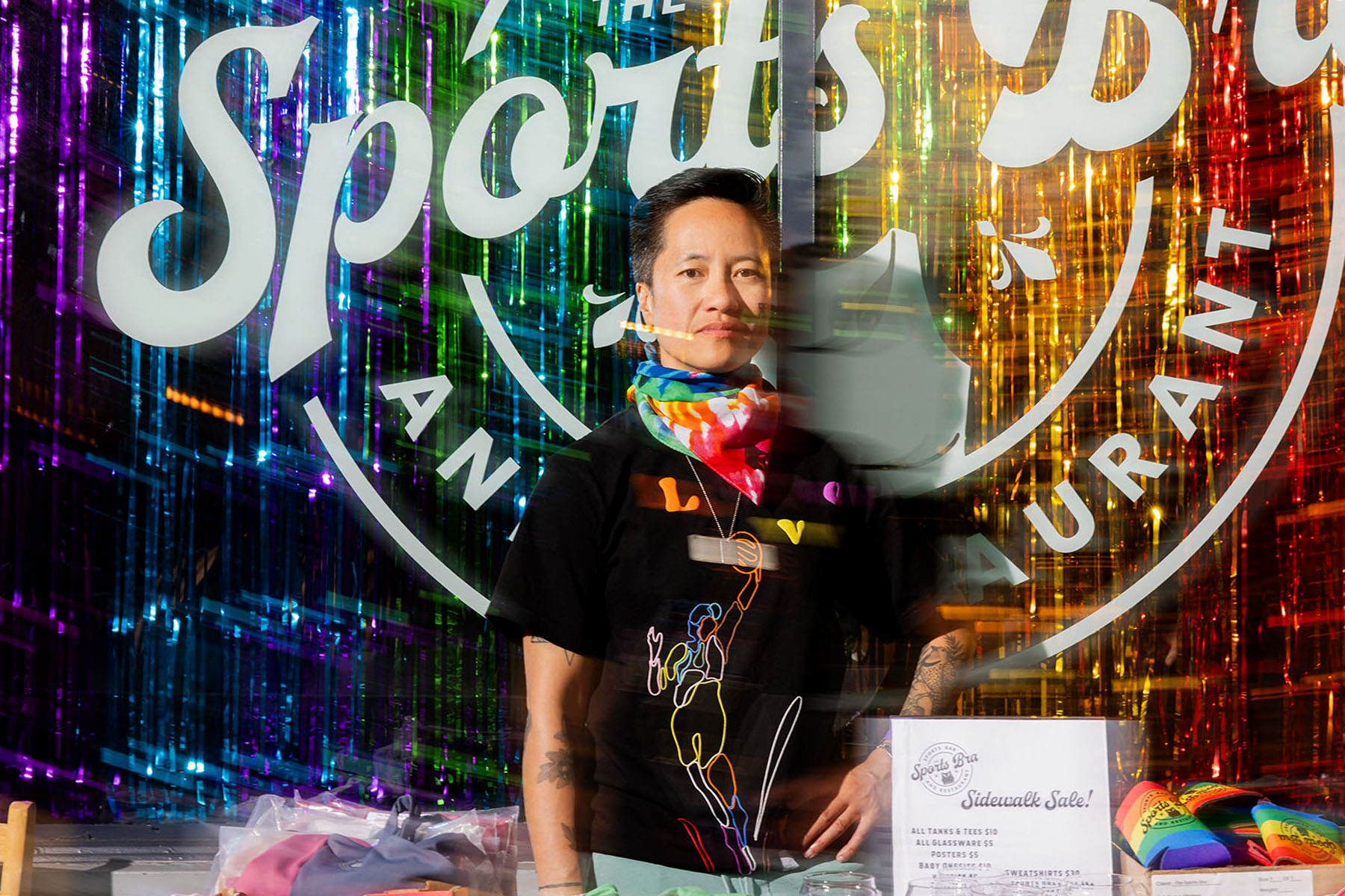 A portrait of Jenny Nguyen in front of her bar, The Sports Bra. The window is decorated with rainbow colored tassels.