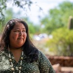 Gabriella Cázares-Kelly, wearing a green patterned blouse and a beaded necklace, stands outdoors. She looks off to the side with a thoughtful expression. Trees and greenery are visible in the blurred background.