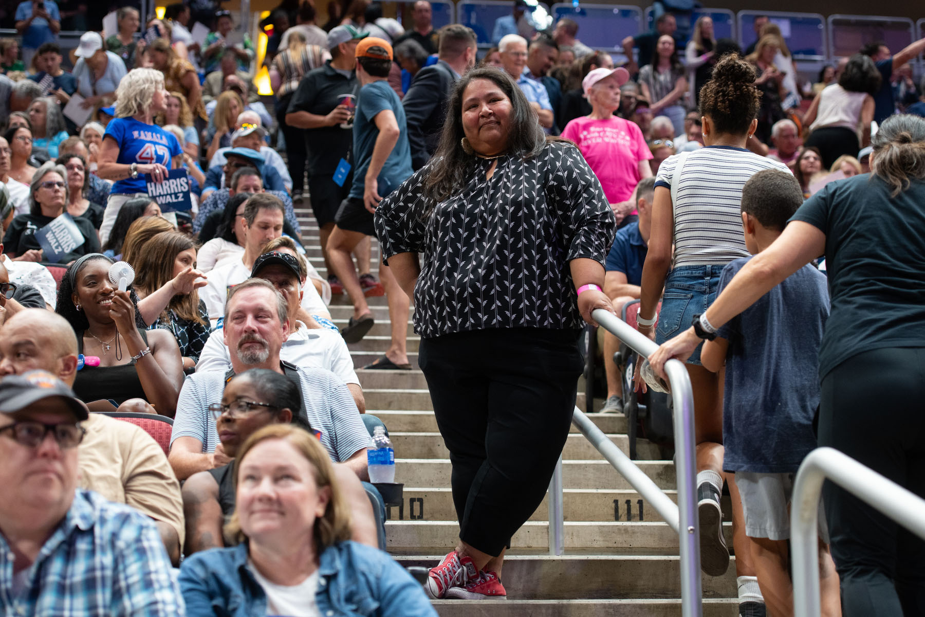 Gabriella Cázares-Kelly stands on the steps inside an arena filled with people attending a campaign rally for Vice President Kamala Harris. She wears a black patterned top and smiles, while attendees around her are engaged in conversations or looking around.