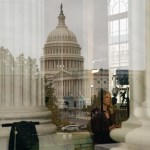 A woman stands near a column, with a reflection of the Capitol in the background.