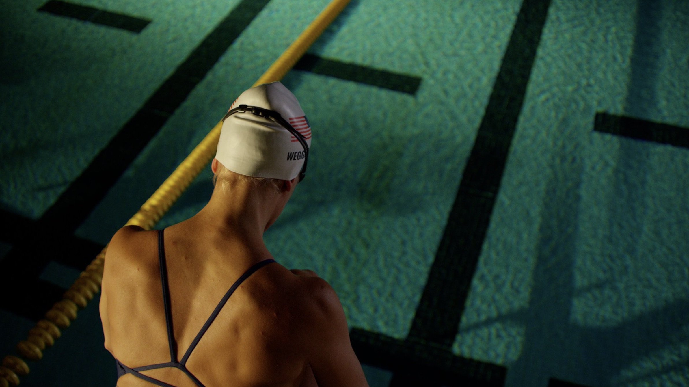 A close-up of Mallory Weggemann wearing a swim cap with the name “Weggemann” printed on it and a USA flag. The swimmer's back is turned to the camera, positioned at the edge of a swimming pool lane.