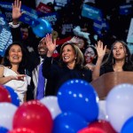 Vice President Kamala Harris is seen smiling and waving from a stage, surrounded by her family and extended family who are also waving and cheering.