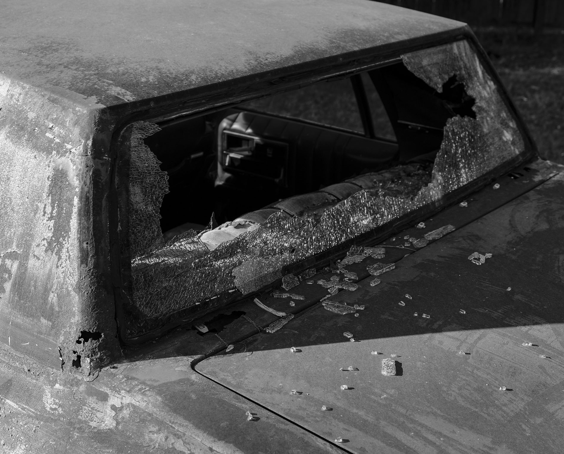 A black-and-white close-up photograph of a car with a broken rear windshield. The car's back window is shattered, and glass fragments are scattered across the trunk and interior.