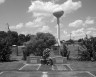 A black-and-white photograph of a cemetery with two gravestones in the foreground. A tall water tower is visible in the background against a partly cloudy sky. The gravestones are adorned with flowers, and a fence encloses the area.