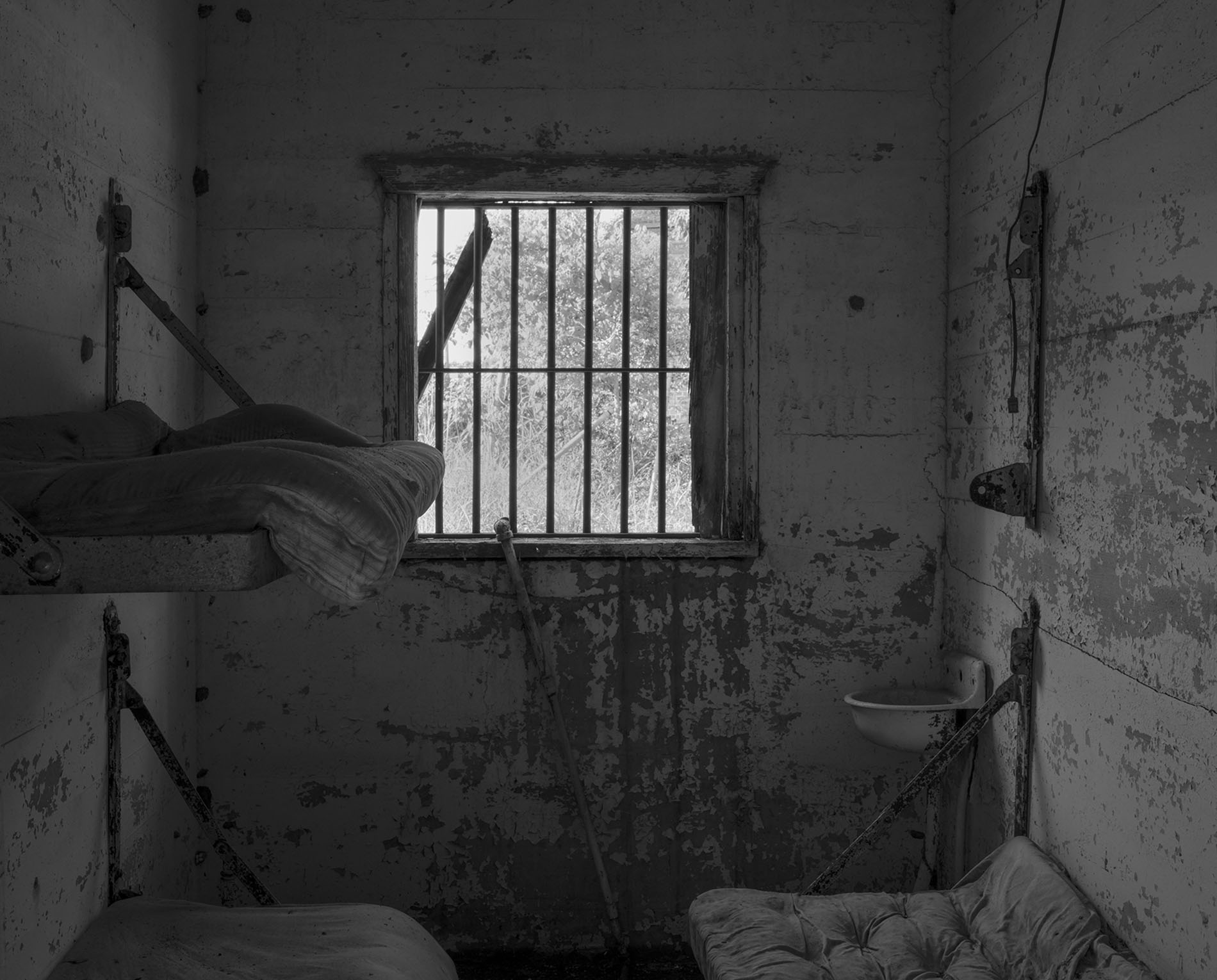 A black-and-white photograph of an old, deteriorated prison cell. The cell has peeling paint on the walls, a small barred window in the center with light filtering through, and two metal-framed bunk beds with worn mattresses. A small, rusted sink is mounted on the wall to the right, and a broken rod or pipe leans against the wall below the window.