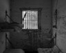 A black-and-white photograph of an old, deteriorated prison cell. The cell has peeling paint on the walls, a small barred window in the center with light filtering through, and two metal-framed bunk beds with worn mattresses. A small, rusted sink is mounted on the wall to the right, and a broken rod or pipe leans against the wall below the window.