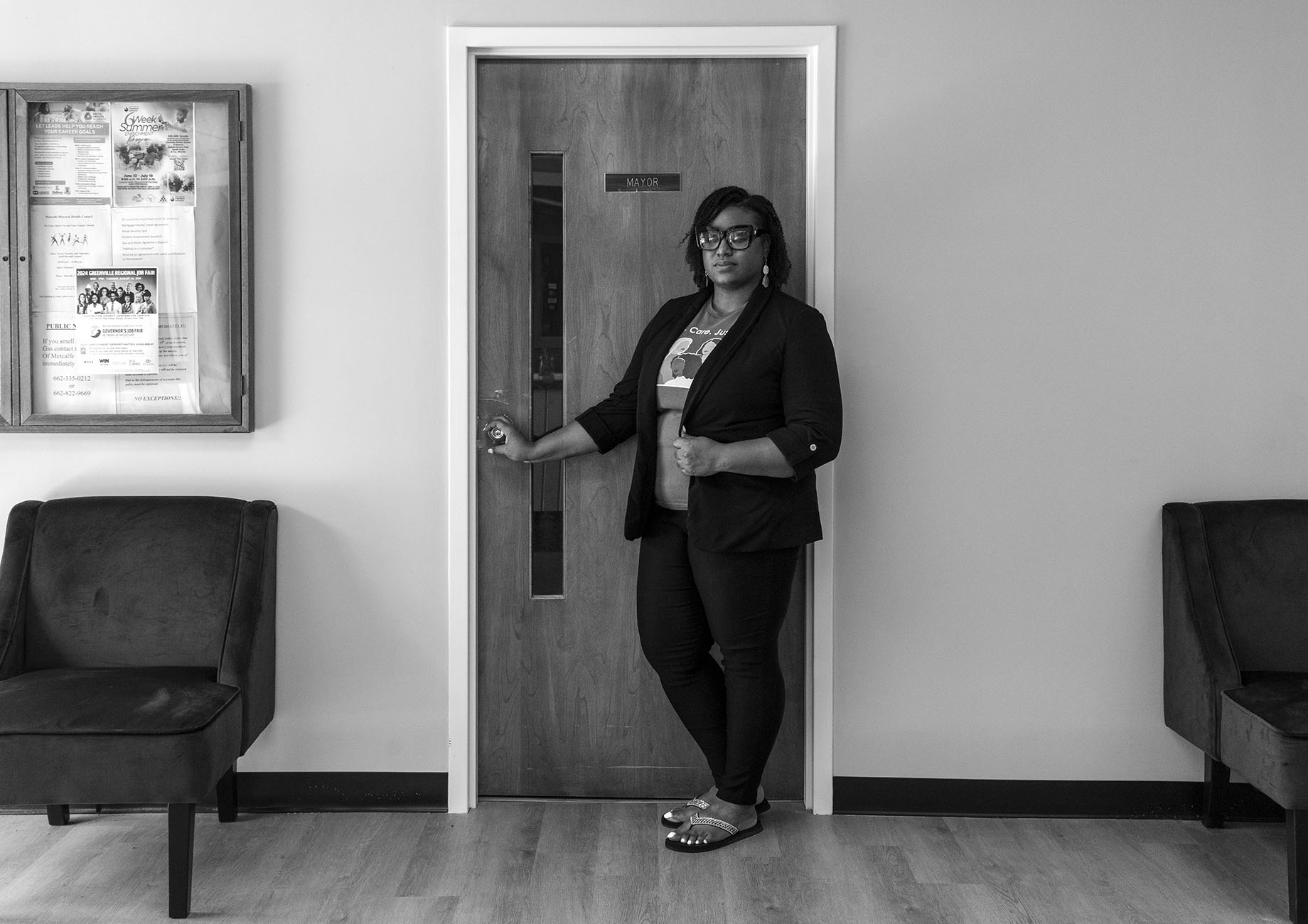 A black-and-white photograph of a woman standing in front of a door labeled "Mayor." She is wearing a blazer and glasses, and she is holding the door handle while looking directly at the camera.