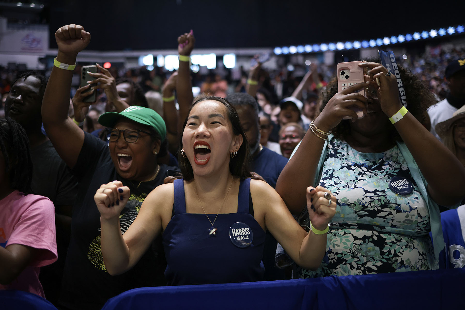 A diverse group of excited supporters cheer and raise their fists at a Harris campaign rally. In the foreground, a woman wearing a blue dress and a "Harris Walz" button shouts with joy, while another woman to her right takes a photo with her phone.