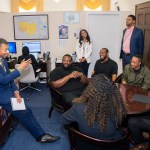 Several people sit and stand together, talking around a table in an office.