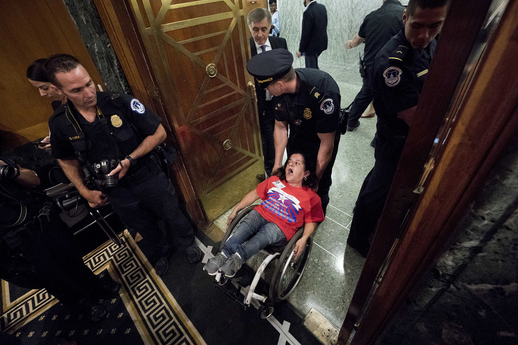 Colleen Flanagan wears a red shirt as she is being forcibly removed from the Capitol building by a police officer who is holding the back of her wheelchair. She appears to be shouting. Several officers are nearby, one of them holding a camera.