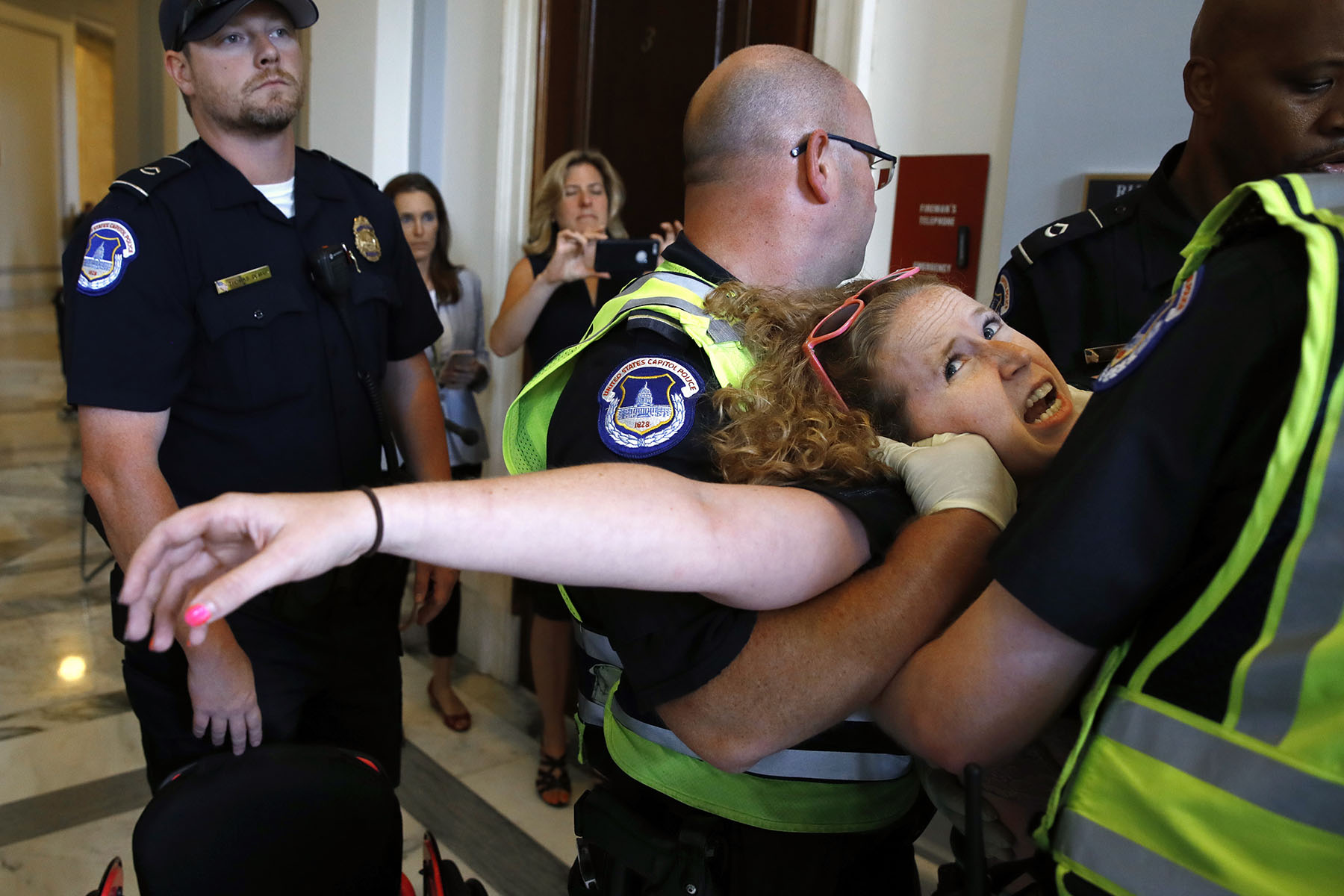 Stephanie Woodward is being restrained by police officers in a hallway. One officer holds her arm while another wraps his arm around her neck.