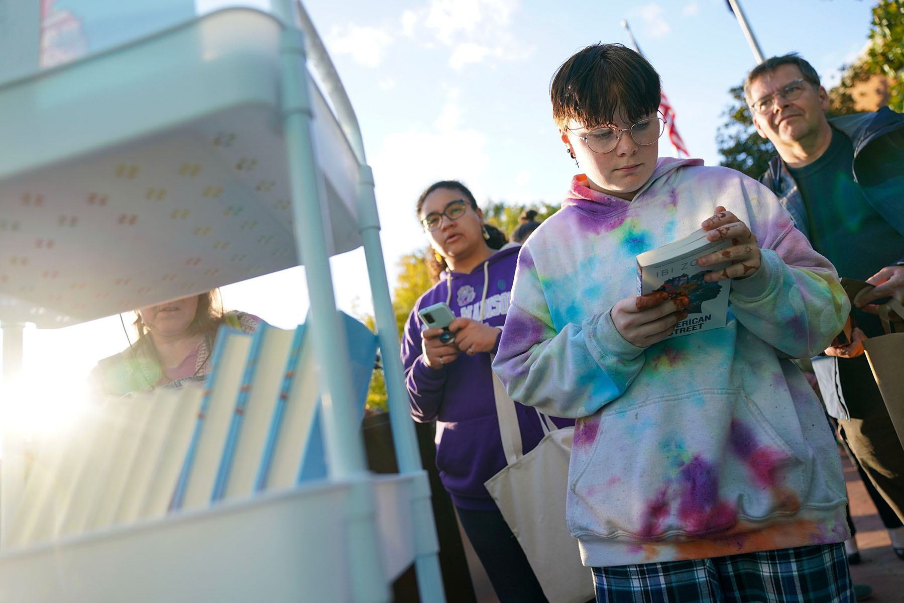 People read banned books outdoors while standing near a book cart.