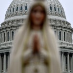 A blurred statue of the Virgin Mary stands in the foreground, with the U.S. Capitol building dome visible in the background.