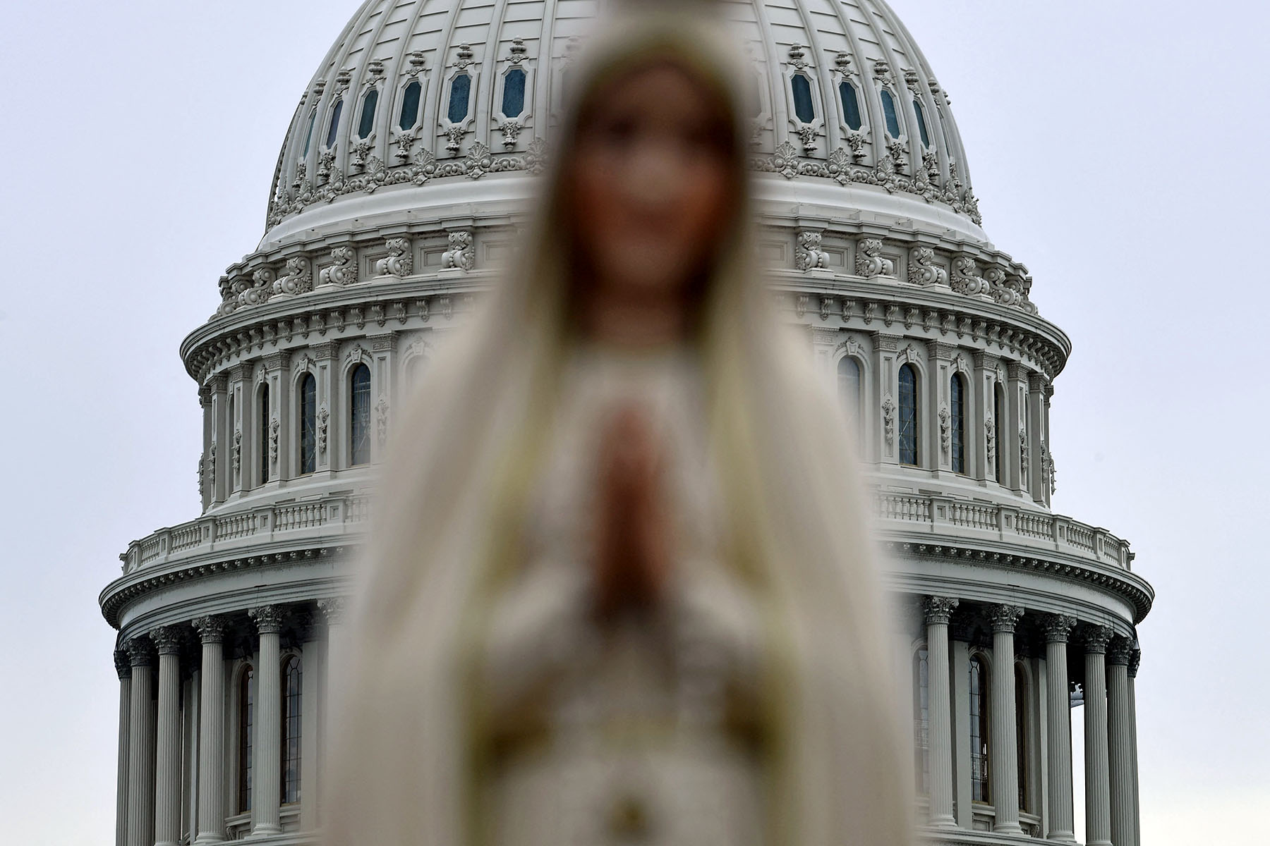 A blurred statue of the Virgin Mary stands in the foreground, with the U.S. Capitol building dome visible in the background.