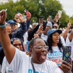 A group of people raising their hands and holding up cell phones during the Crime Survivors Speak March in Washington, D.C. Many are wearing white shirts with slogans like 