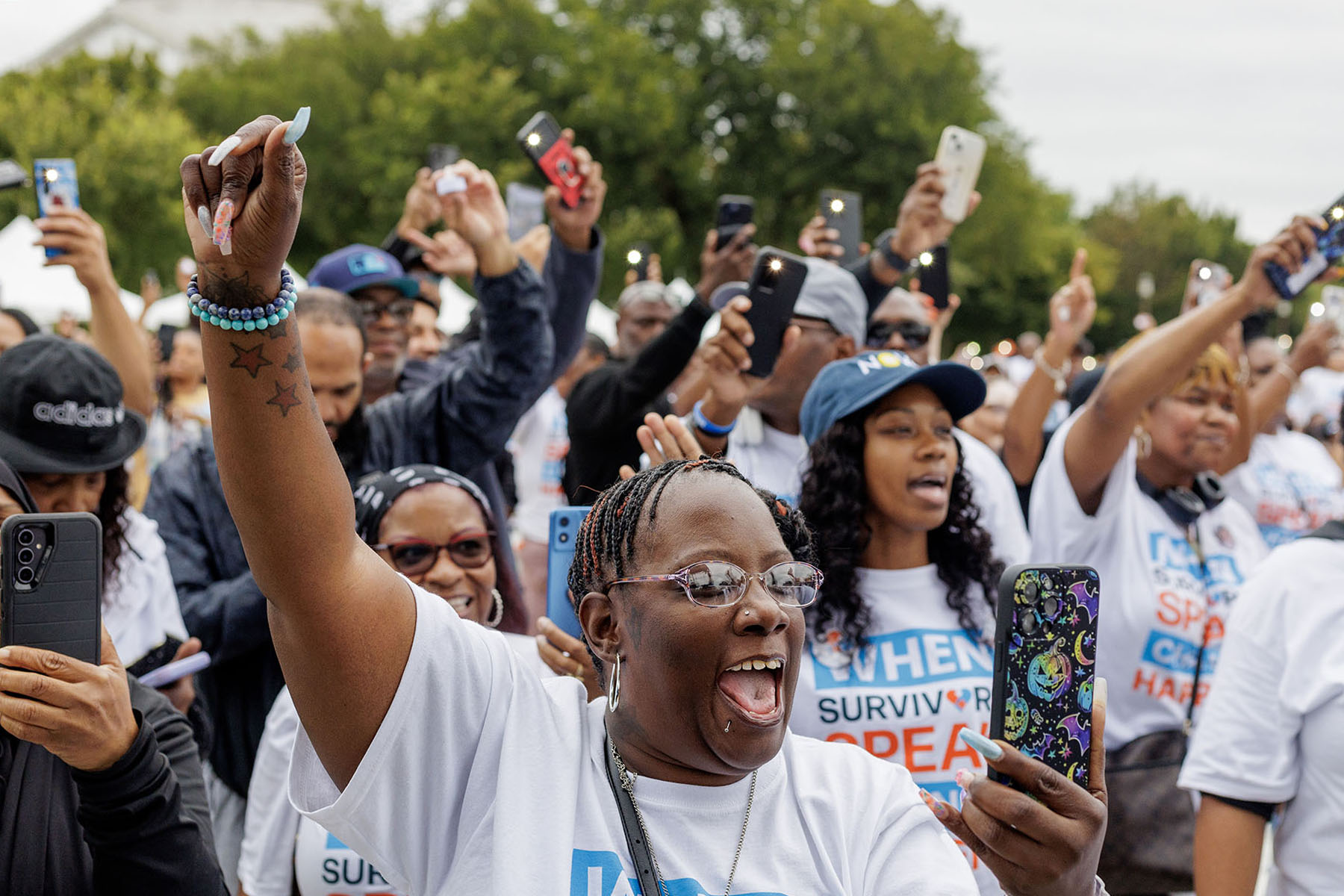 A group of people raising their hands and holding up cell phones during the Crime Survivors Speak March in Washington, D.C. Many are wearing white shirts with slogans like "When Survivors Speak" and are smiling and cheering.