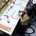 A woman using a wheelchair works at a drafting table, drawing with a ruler surrounded by various tools and materials in a workshop.