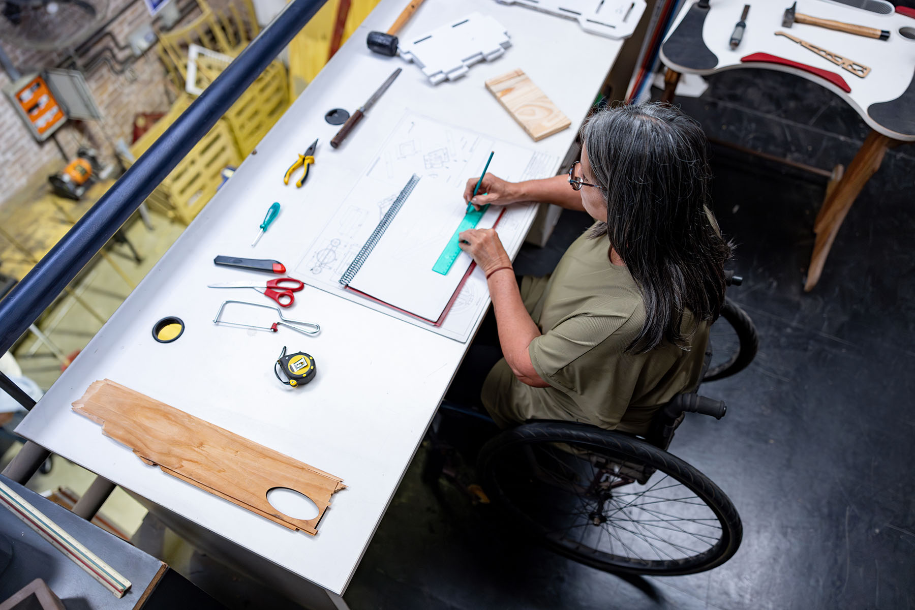 A woman using a wheelchair works at a drafting table, drawing with a ruler surrounded by various tools and materials in a workshop.