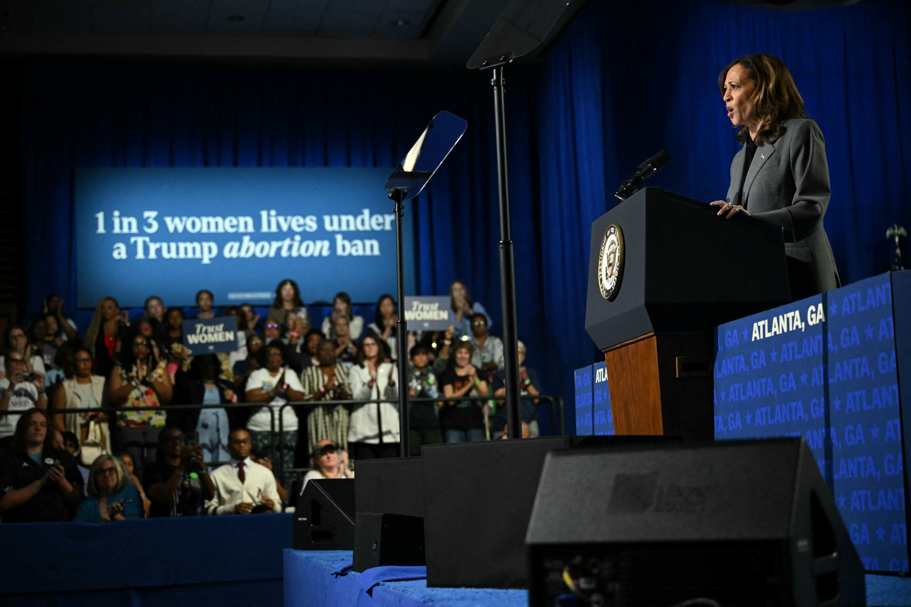 Vice President Kamala Harris stands at a lectern near a crowd sitting under a sign that says "1 in 3 women lives under a Trump abortion ban"