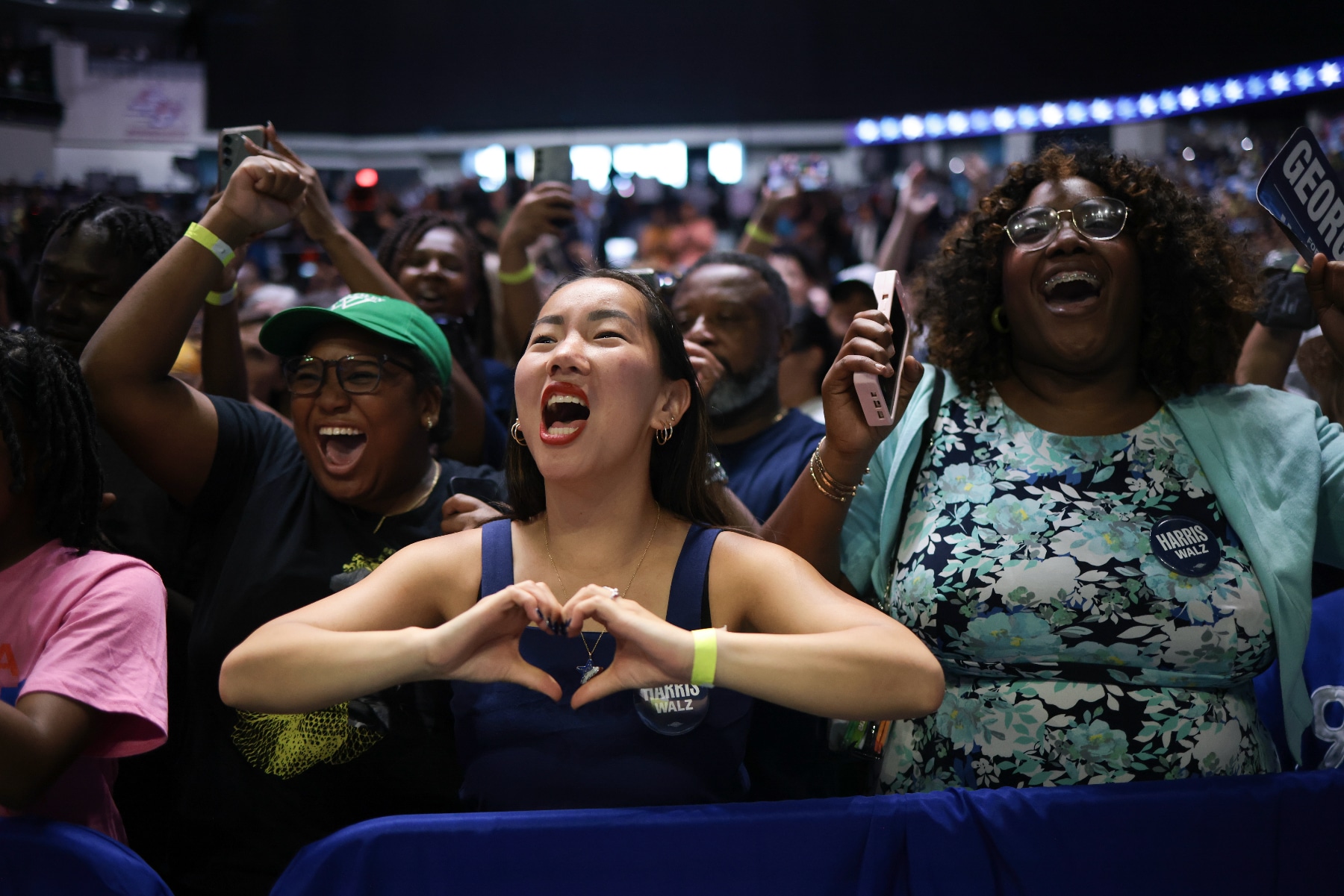 People cheer for Kamala Harris at a campaign event, with one person making a heart with her hands as she cheers