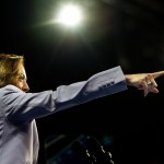 Vice President Kamala Harris, seen from the side, points forward with her right arm during a rally. A bright light shines above her head as she addresses the crowd.