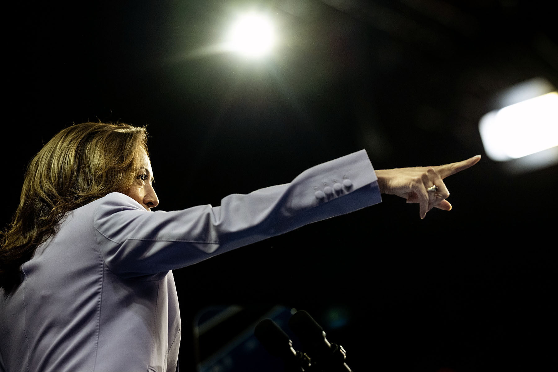 Vice President Kamala Harris, seen from the side, points forward with her right arm during a rally. A bright light shines above her head as she addresses the crowd.