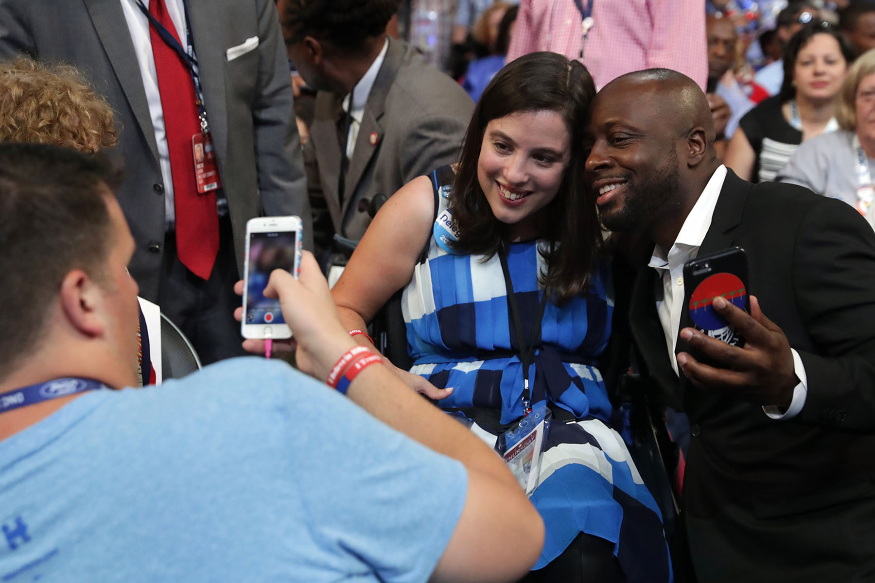 Anastasia Somoza, an international disability rights advocate, smiles as she poses for a photo with a man at the Democratic National Convention. She is seated in a wheelchair, wearing a blue and white dress, while another person holds up a phone to take the picture.