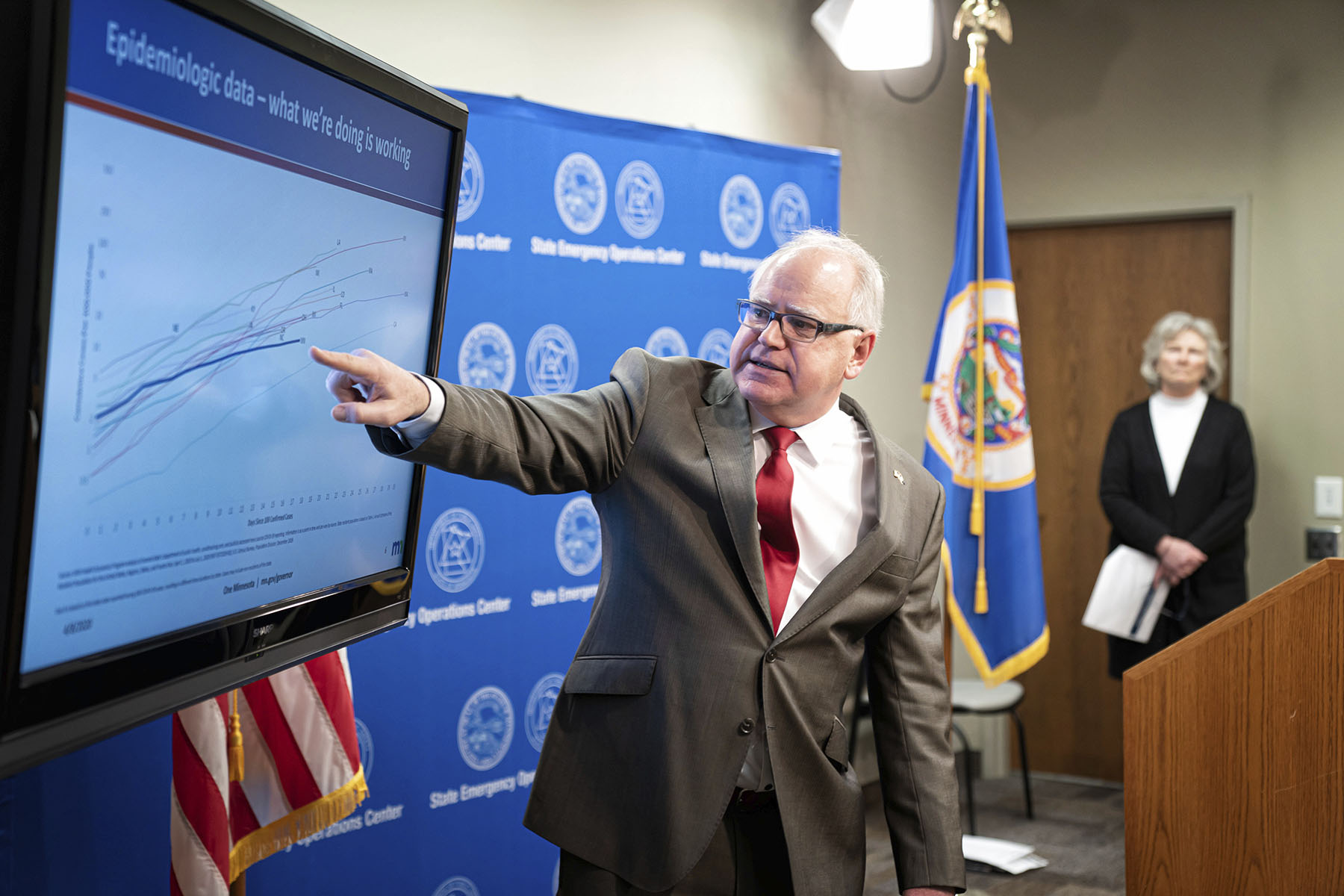 Minnesota Governor Tim Walz points to a large screen displaying epidemiologic data during a COVID-19 press conference. He stands in front of a blue background with state emblems, wearing a suit and red tie.