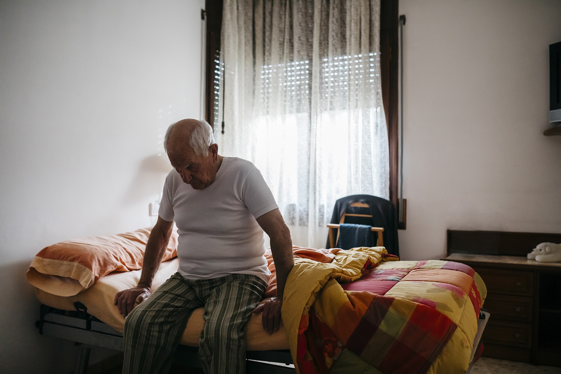 An elderly man sits at the edge of a bed alone in a room with a window at his back.