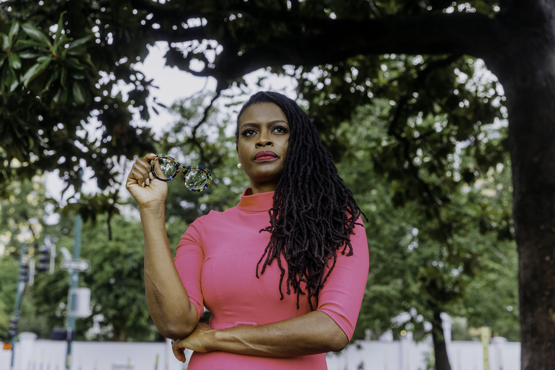 Cassandra Welchlin holding glasses in one hand, standing under a tree with a determined expression. She is wearing a bright pink dress, with her long locs draped over her shoulder, and the background features a park setting.