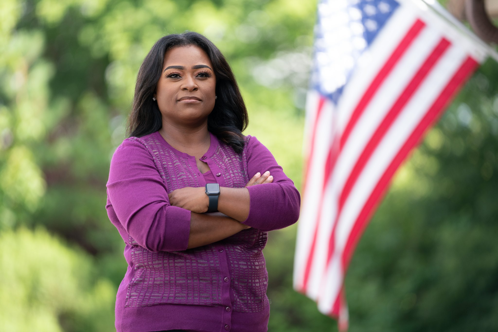 Diamond Staton-Williams, wearing a purple sweater, stands with arms crossed in front of an American flag.